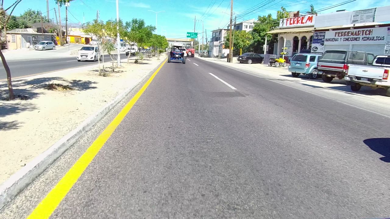 Vehicle lane view on a sunny street with parked cars and storefronts.