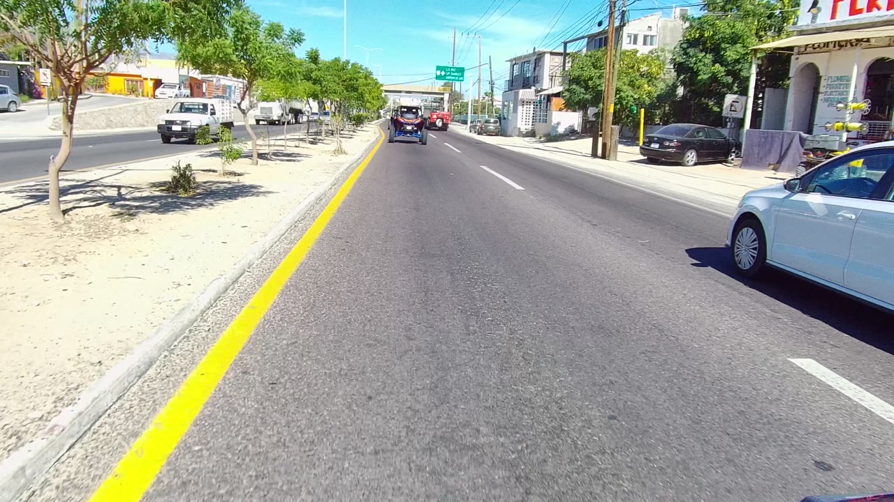 At 0:02, the Los Cabos street view includes a white car entering along the right edge, shops and parked vehicles on the roadside, trees casting small shadows, and no visible people.