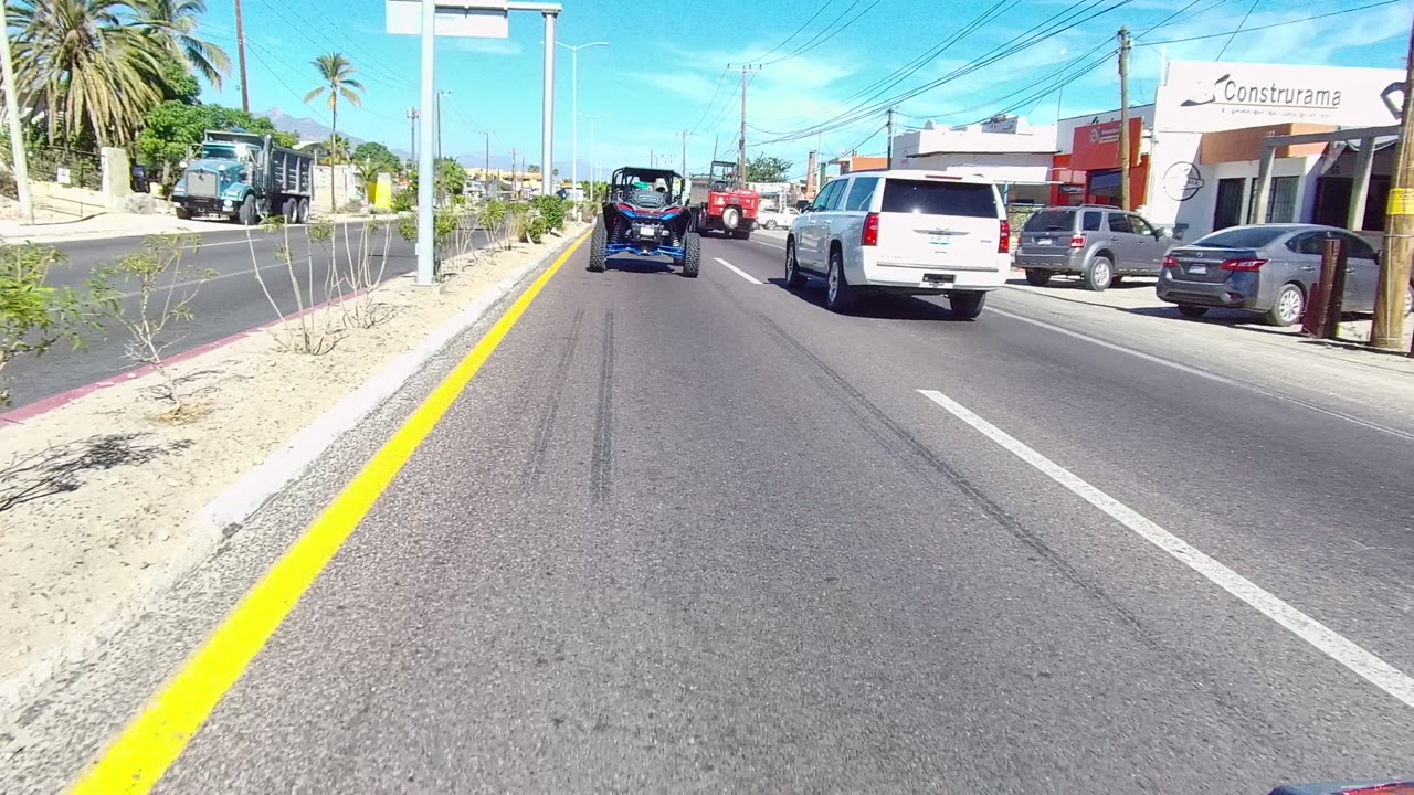 A Los Cabos road view shows a dark truck, a blue off-road vehicle, and a white SUV moving past storefronts and sandy roadside planting, with no visible people in this frame.