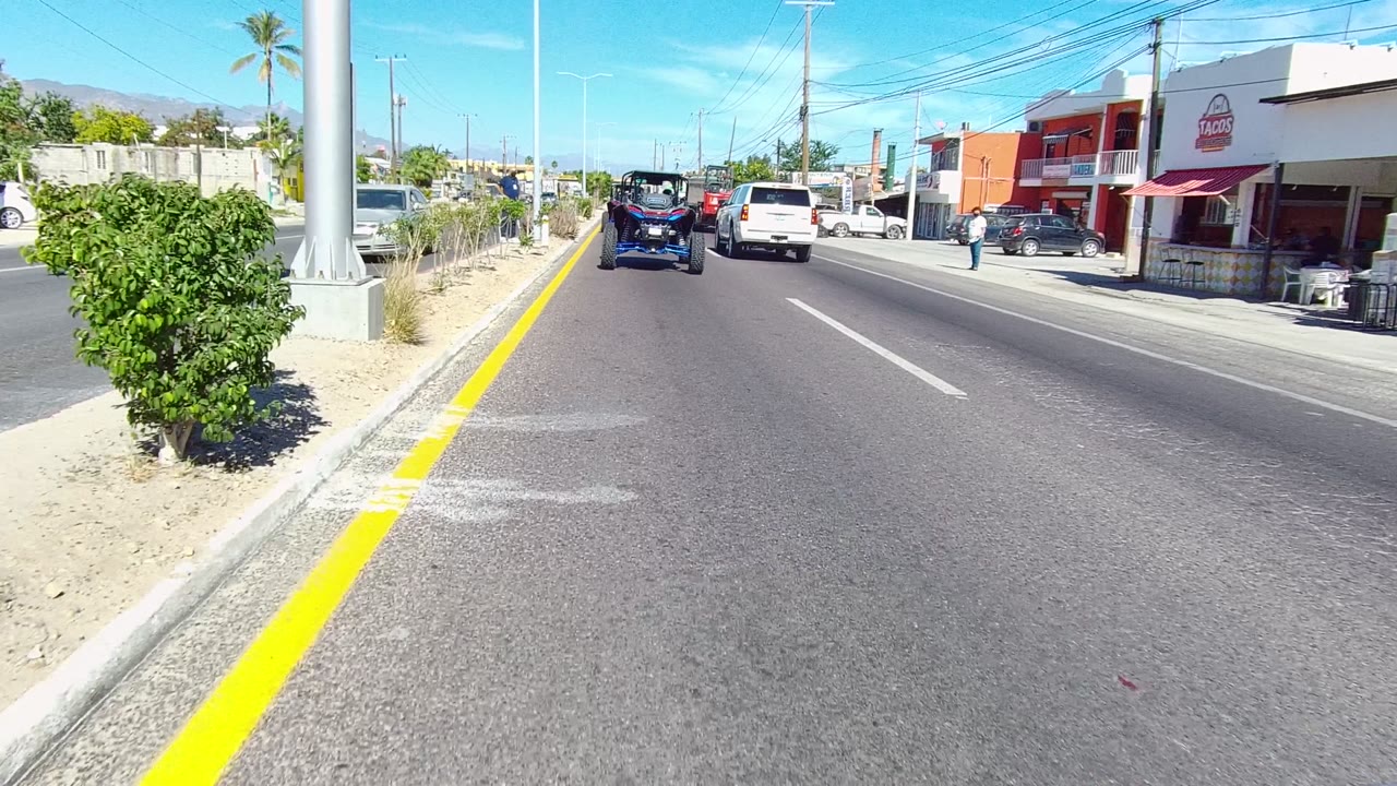 A Los Cabos street frame shows the road opening ahead with traffic, a white SUV, low storefronts, a large green shrub at the median edge, palms, and distant hills.