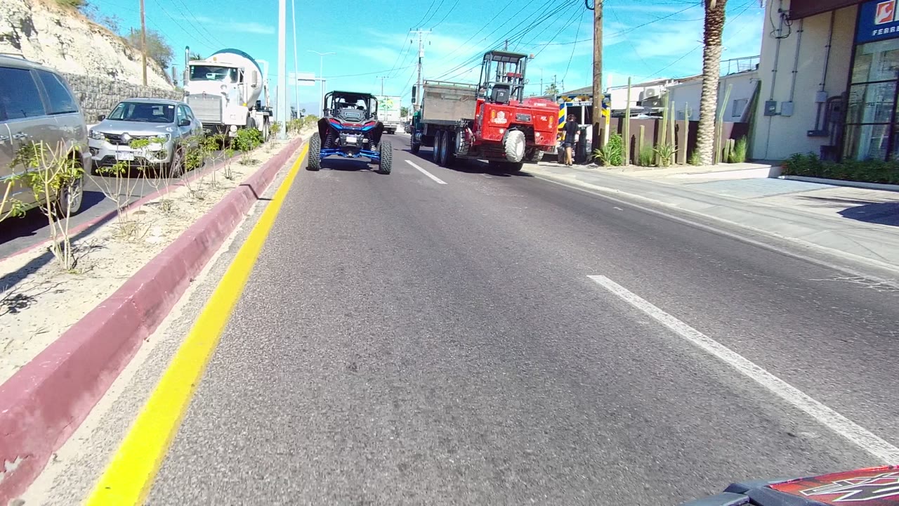 Opening view on a paved Los Cabos road with a yellow curb line at left, parked cars along the roadside, a blue off-road vehicle ahead, a red truck or tractor farther forward, and no visible people.