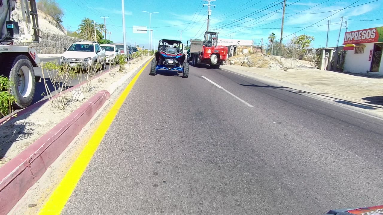 Mid-clip Los Cabos road frame with the blue off-road vehicle and red truck ahead, parked cars along both sides, roadside shops, a rocky hillside, overhead wires, palms, and no visible people.