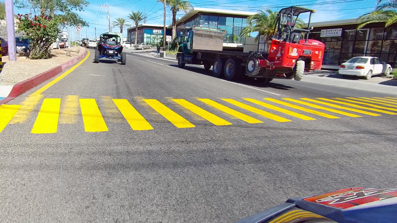 A red dump truck and small blue off-road vehicle travel along a sunny Los Cabos street with yellow crosswalk markings, parked cars, glass storefronts, palm trees, and no visible people.