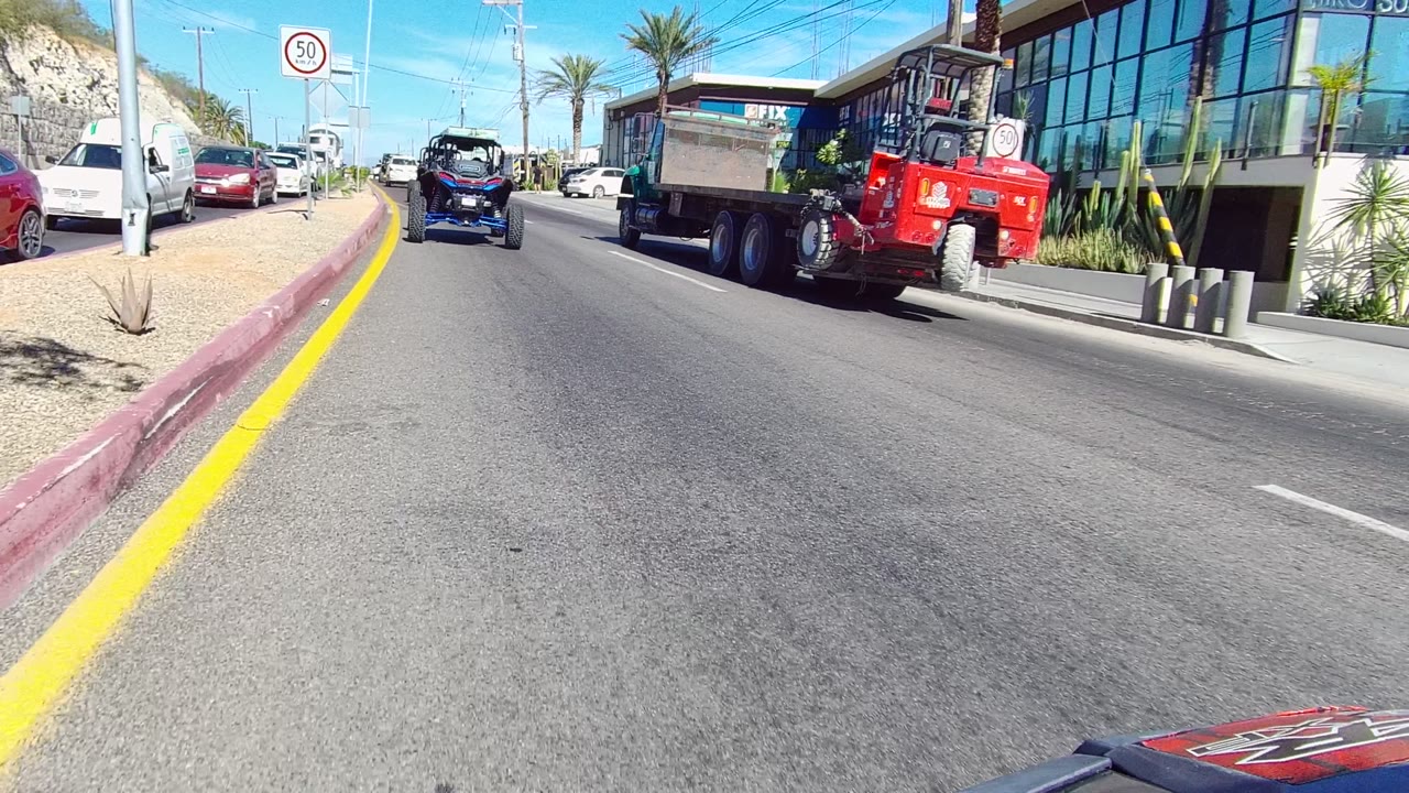 A red dump truck dominates the right side of a Los Cabos street scene, with a small blue vehicle ahead, a white van by the curb, glass storefronts, palms, and no visible people.