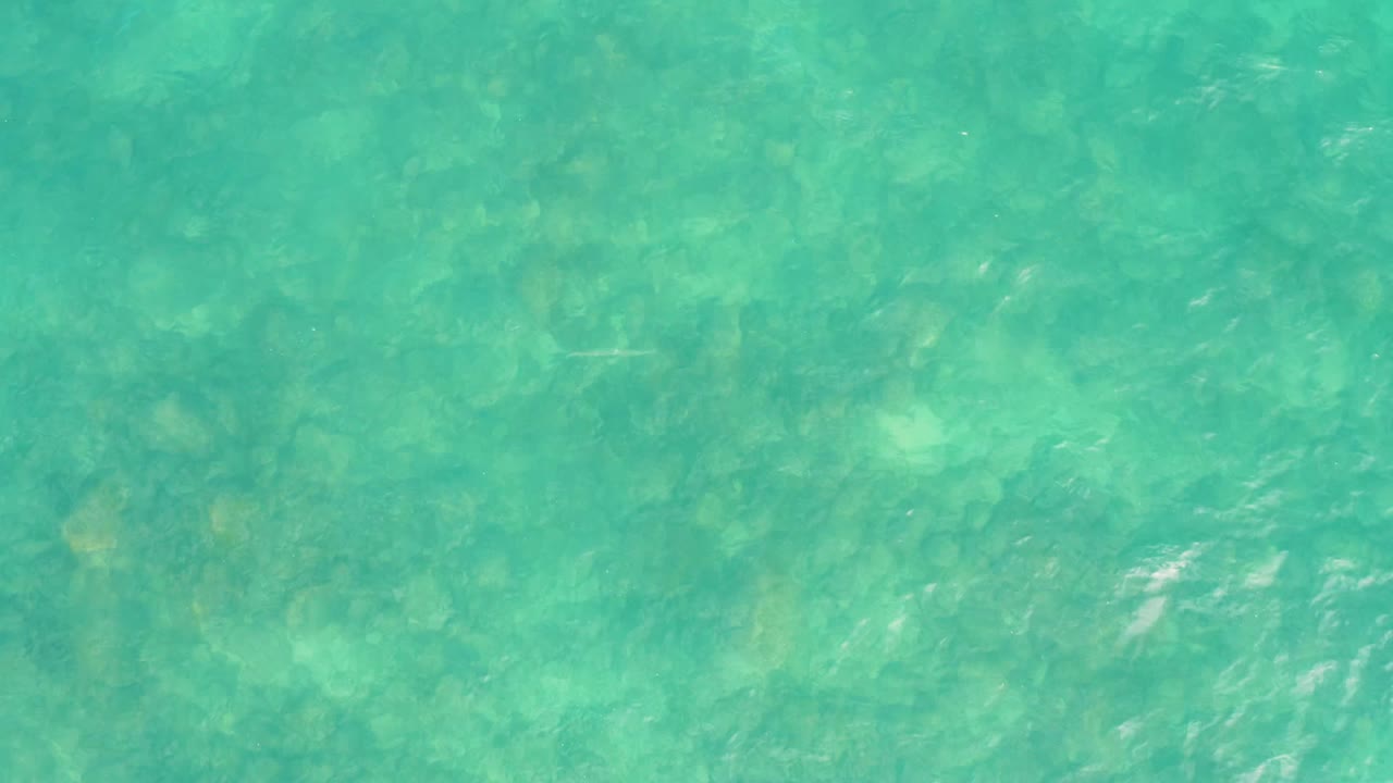 Downward view of turquoise sea at El Tule Los Cabos with faint white ripple streaks and mottled seabed detail below, with no visible people.