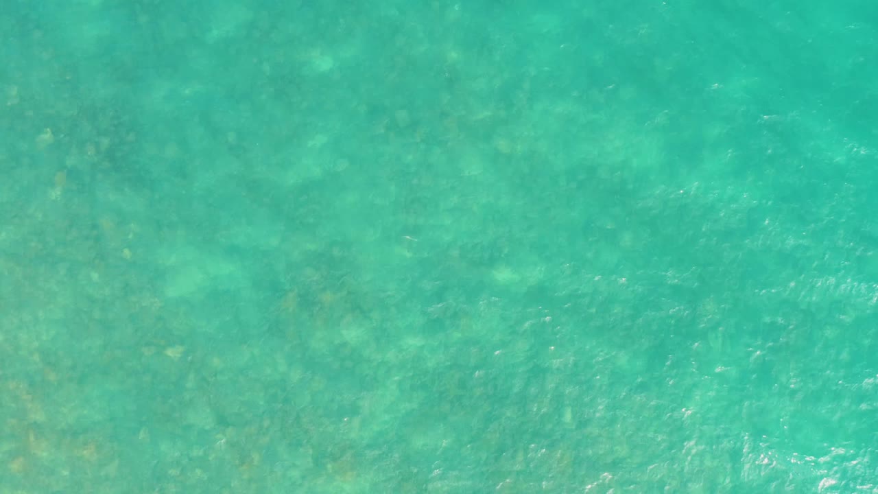 Overhead view of turquoise shallow water at El Tule Los Cabos with darker seabed patches faintly visible and no visible people.