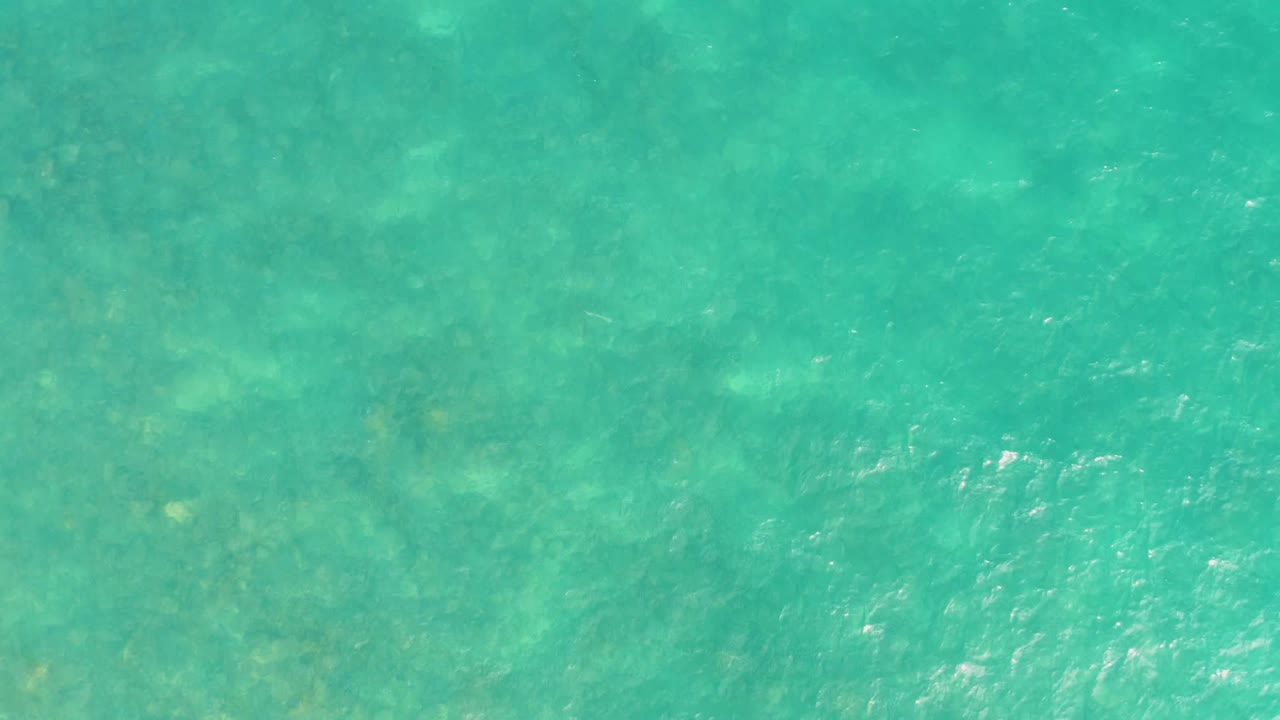 Transparent turquoise sea at El Tule Los Cabos shows mottled darker underwater areas beneath gentle ripples and no visible people.