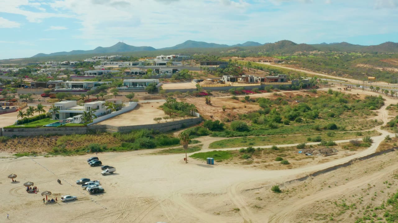 Aerial view in El Tule Los Cabos showing a sandy foreground road with cars, scrubby lots, modern buildings, and distant mountains under a pale blue sky.