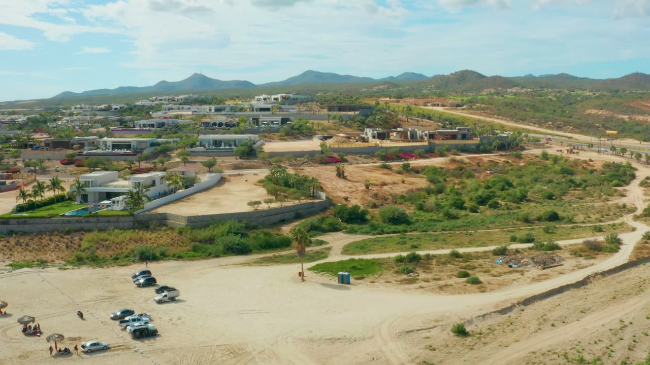 Drone view of cars near a dirt road with low buildings and arid hills beyond.
