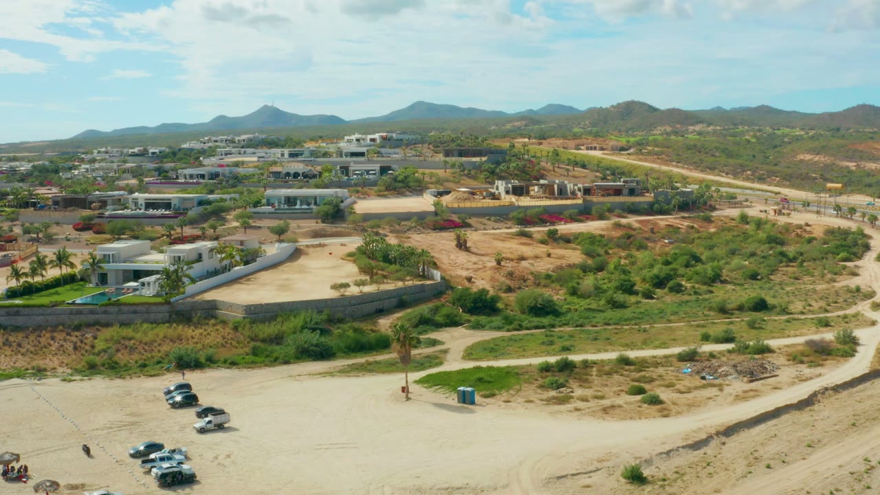 Wide aerial frame in El Tule Los Cabos with the dirt road crossing the lower area, cars clustered near the foreground, scattered desert vegetation, residential-looking buildings, and mountains along the horizon.