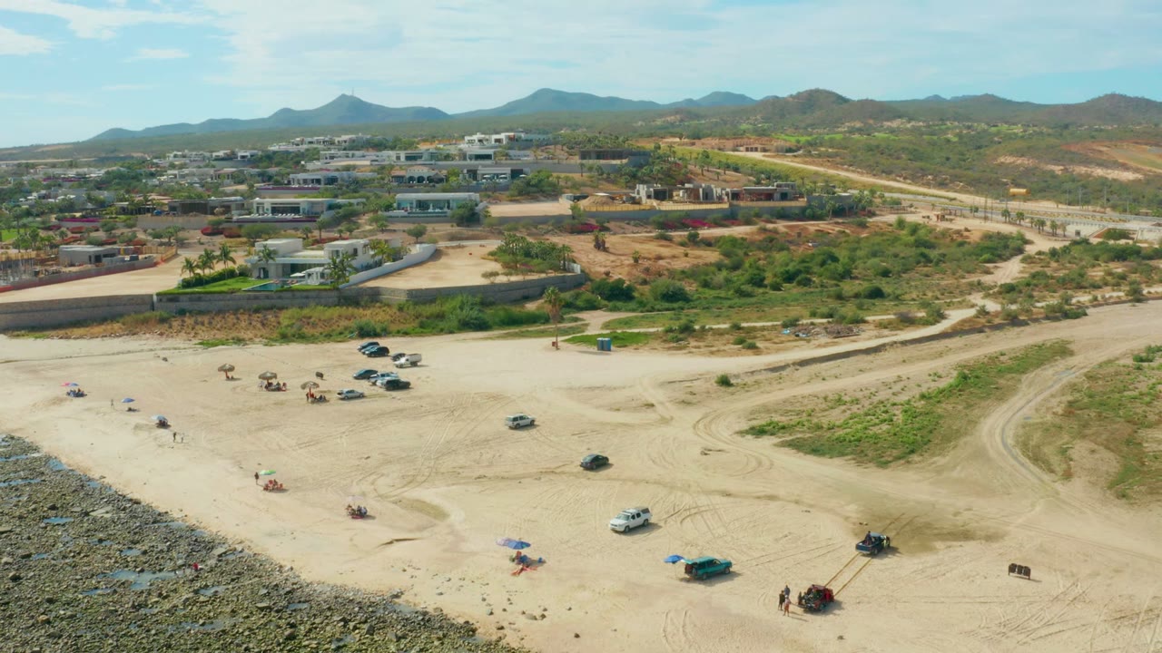 Aerial frame over El Tule Los Cabos showing a sandy beach clearing with scattered vehicles, small umbrellas, people, low buildings, a road, green scrub, and hills under blue sky.