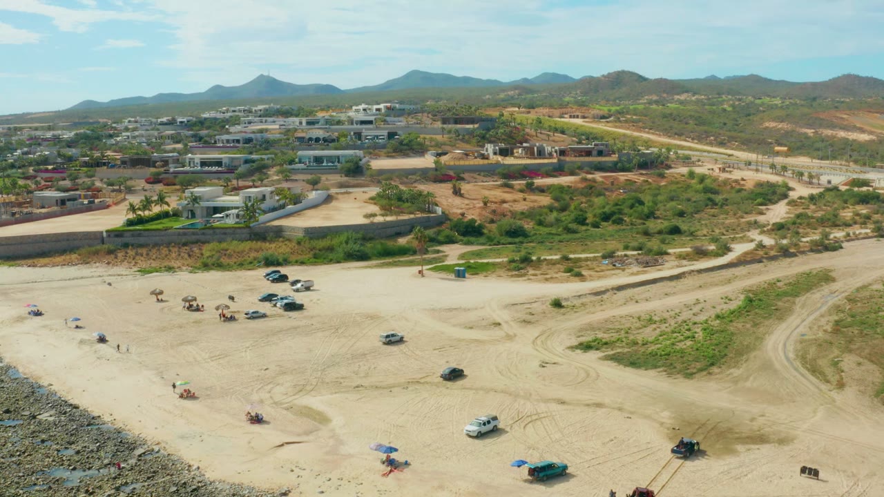 Wide drone view of beach access area with fewer vehicles, umbrellas, road, buildings, and hills.