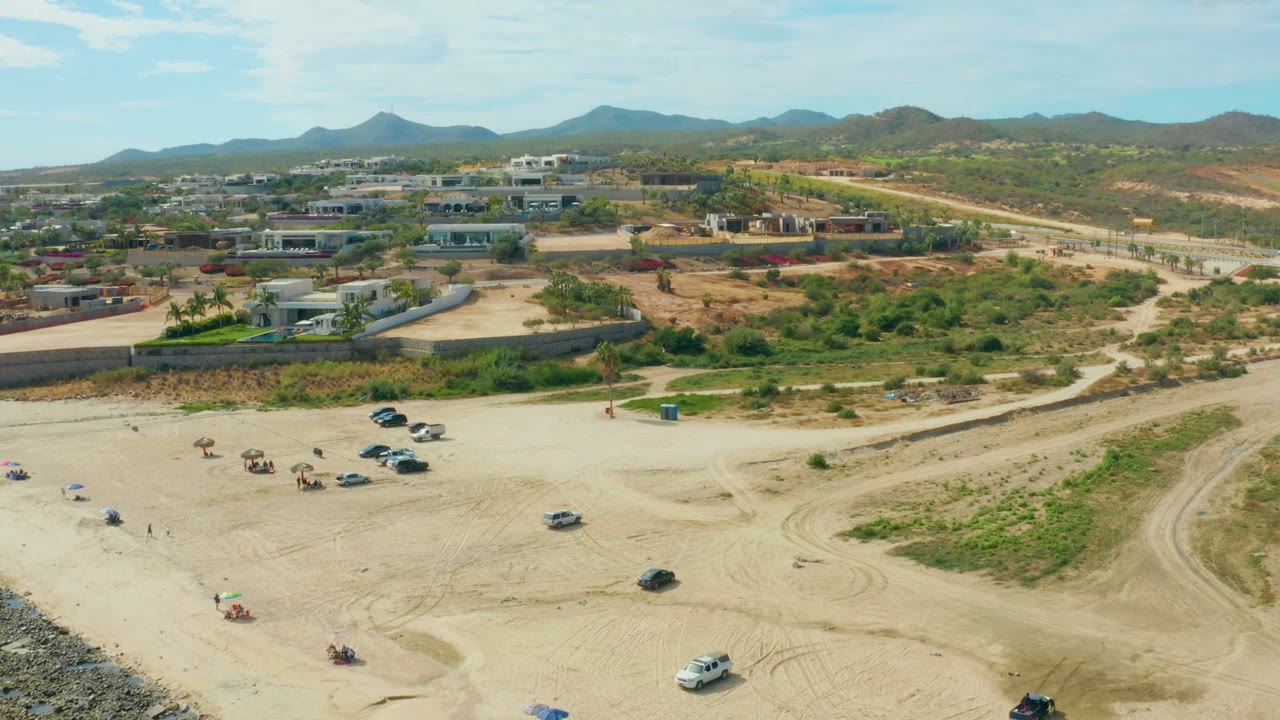 Aerial view in El Tule Los Cabos with scattered cars and umbrellas on pale sand, tiny people visible, a road and buildings beyond, and dry green hills across the horizon.