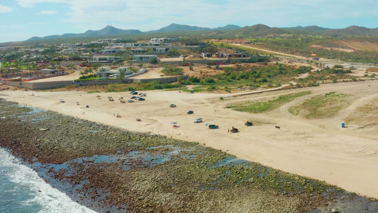 Aerial frame over El Tule Los Cabos showing surf at the lower left, exposed rocky shore, a wide sandy beach with scattered vehicles and beachgoers, and low hills beyond buildings.