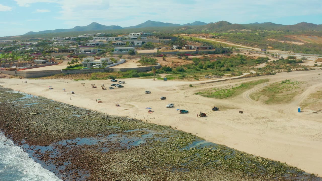 Wide drone view of beach vehicles, small people, rocky tide pools, surf, and hills.
