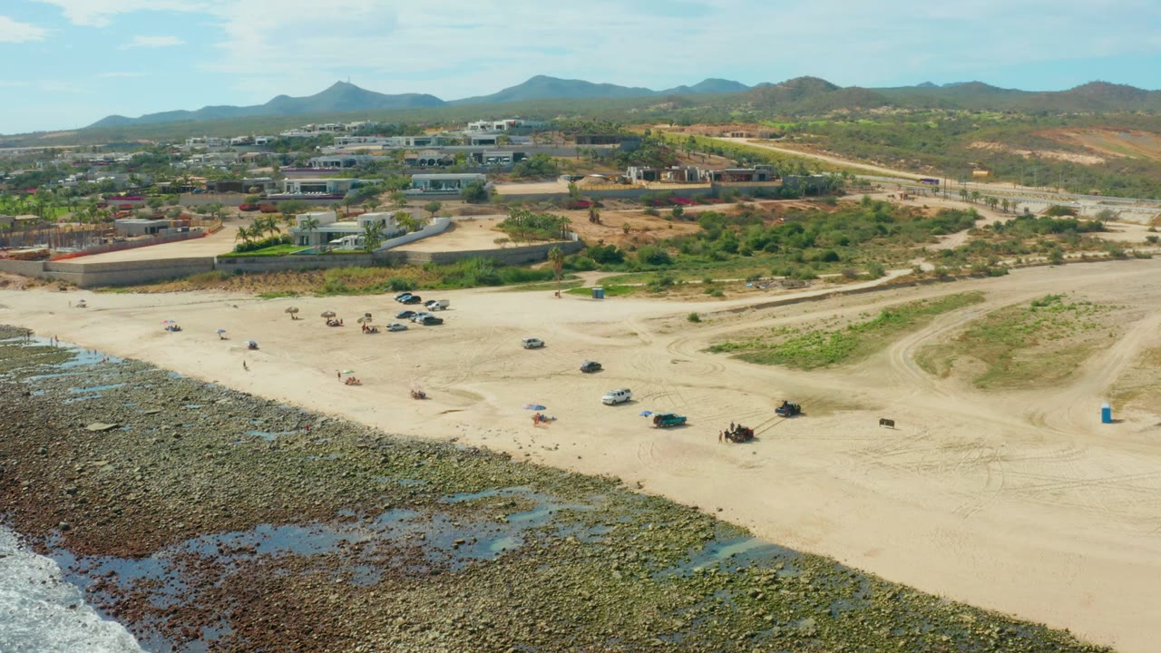 Aerial frame over El Tule Los Cabos where the rocky waterline fills the lower left, cars and small groups sit across the sand, and mountains line the background.