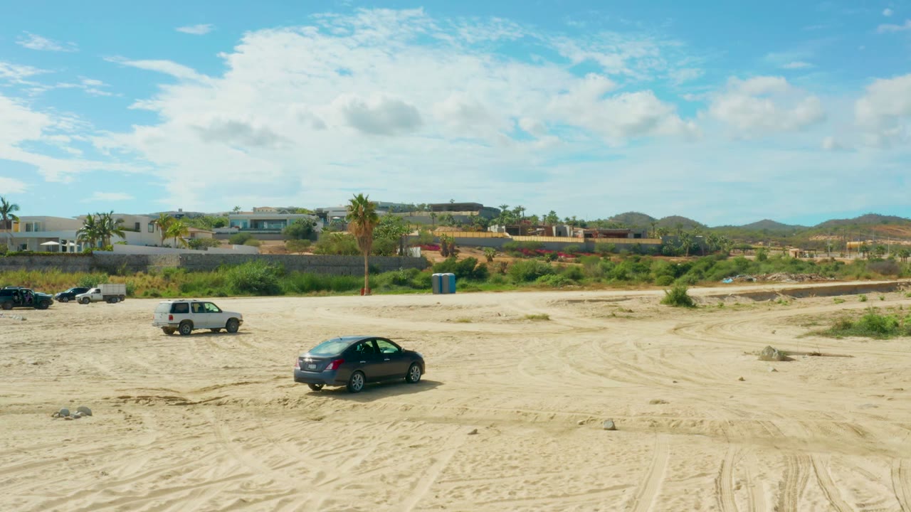 Wide view of a sandy open area in El Tule Los Cabos with parked cars, low buildings, palm trees, distant hills, blue sky, and no visible people.