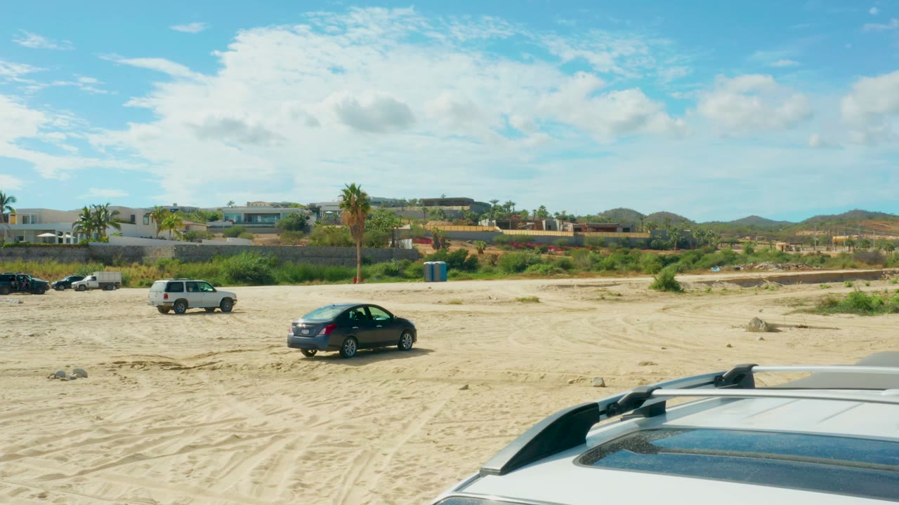 The vehicle roof occupies more of the lower foreground as parked cars sit across sandy ground in El Tule Los Cabos, backed by buildings, palms, low hills, and no visible people.