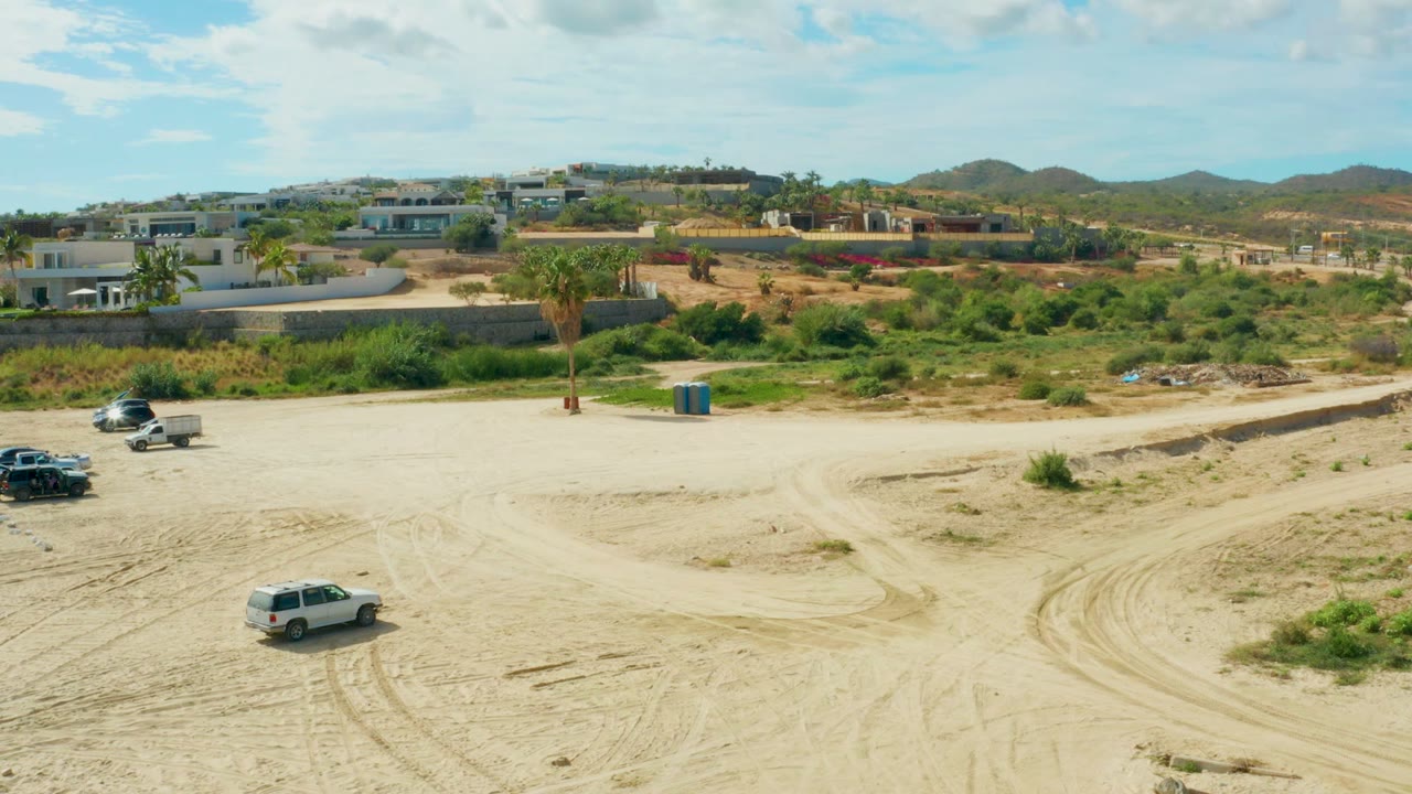 At 0:00 in El Tule Los Cabos, a wide sandy lot shows a white SUV, dark vehicles near the road, low buildings, palms, distant hills, and no visible people.