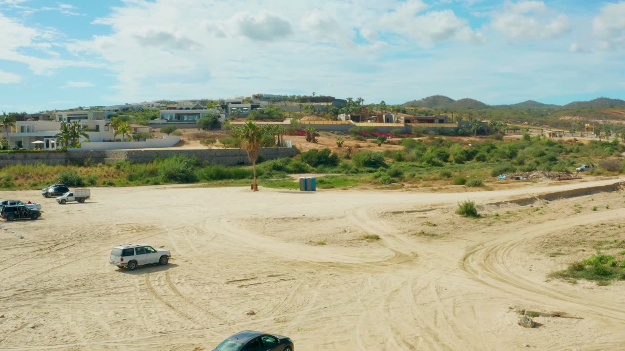 At 0:02 in El Tule Los Cabos, the white SUV crosses the pale dirt foreground as dark vehicles remain nearer the road, with a blue utility structure, low buildings, hills, and no visible people.