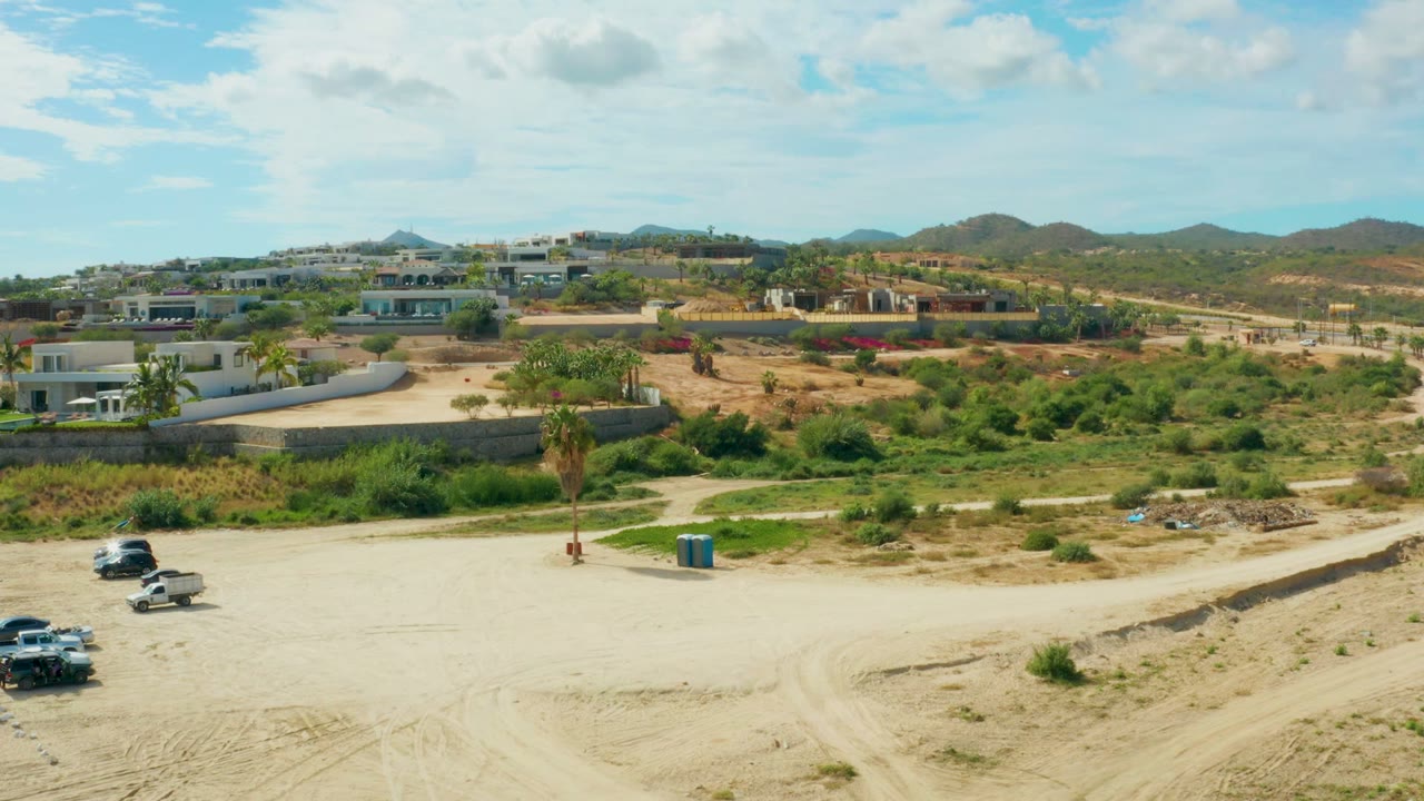 Opening frame of El Tule Los Cabos showing a wide sandy lot with tire tracks, parked vehicles, low buildings, green scrub, distant hills, and no visible people.