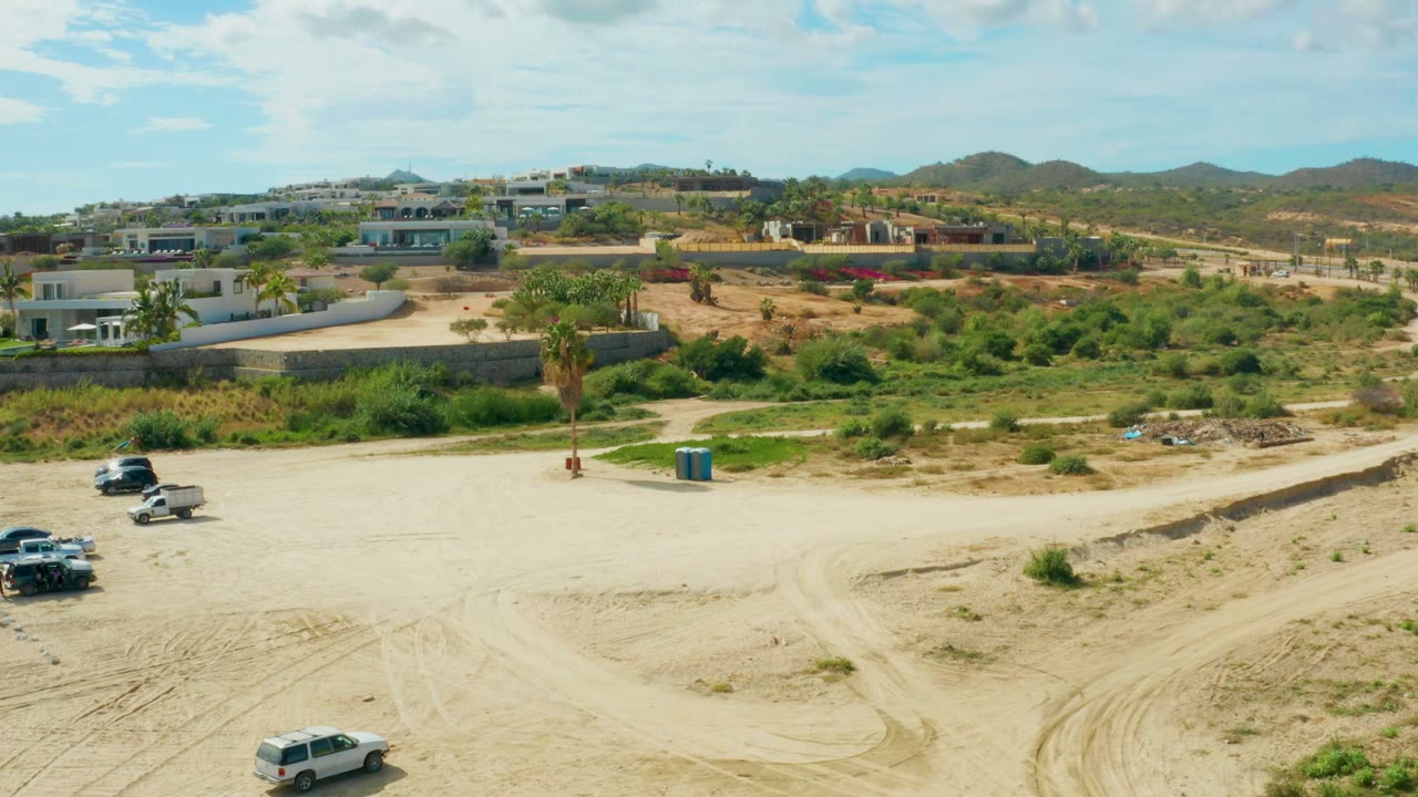 Wide aerial-style view in El Tule Los Cabos as the white SUV moves farther across pale sand with curved tire tracks, scattered vehicles, hillside buildings, and no visible people.
