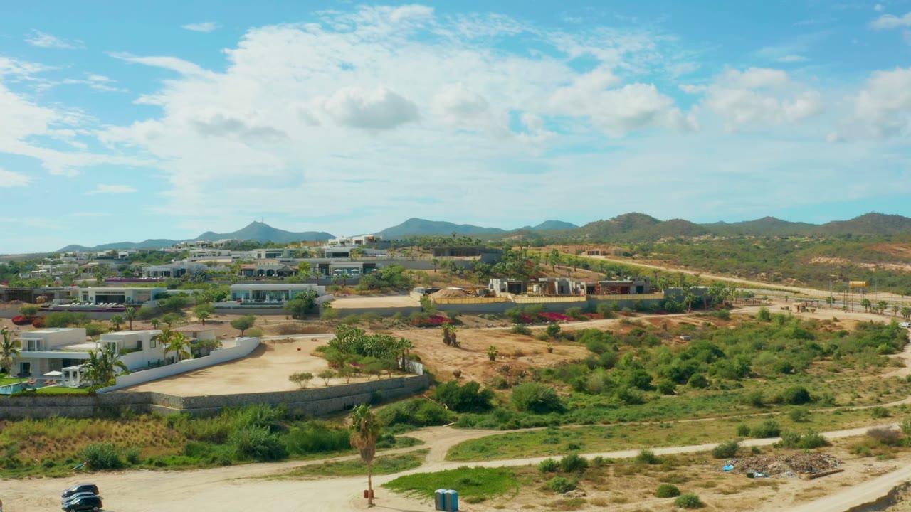 Opening aerial frame in El Tule Los Cabos, Baja California Sur, with a pale dirt road at the bottom, scrubby green terrain, low modern buildings, distant hills, scattered clouds, and no visible people.