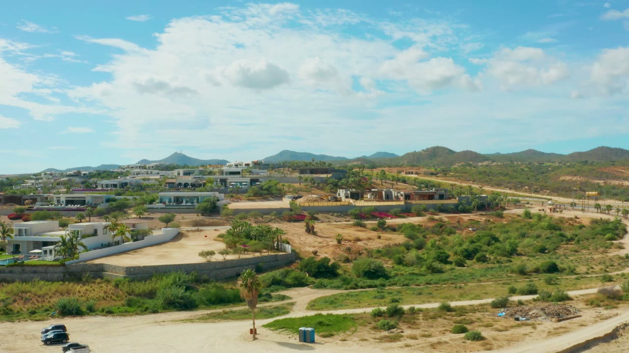 Vehicles travel near the lower edge of a dirt road below buildings and hills.
