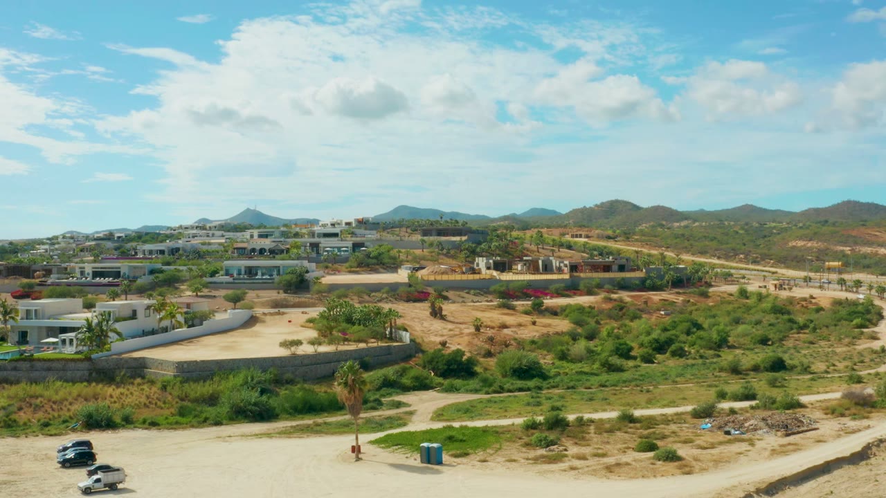 At 0:02 in El Tule Los Cabos, vehicles sit near the lower dirt road, two blue portable toilets stand beside the open ground, and buildings and hills fill the background with no visible people.