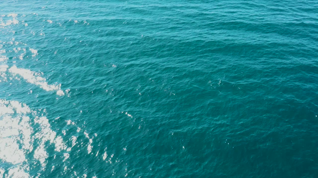Overhead ocean view in El Tule Los Cabos with white foam clustered along the left side, darker turquoise ripples across the right, and no visible people.
