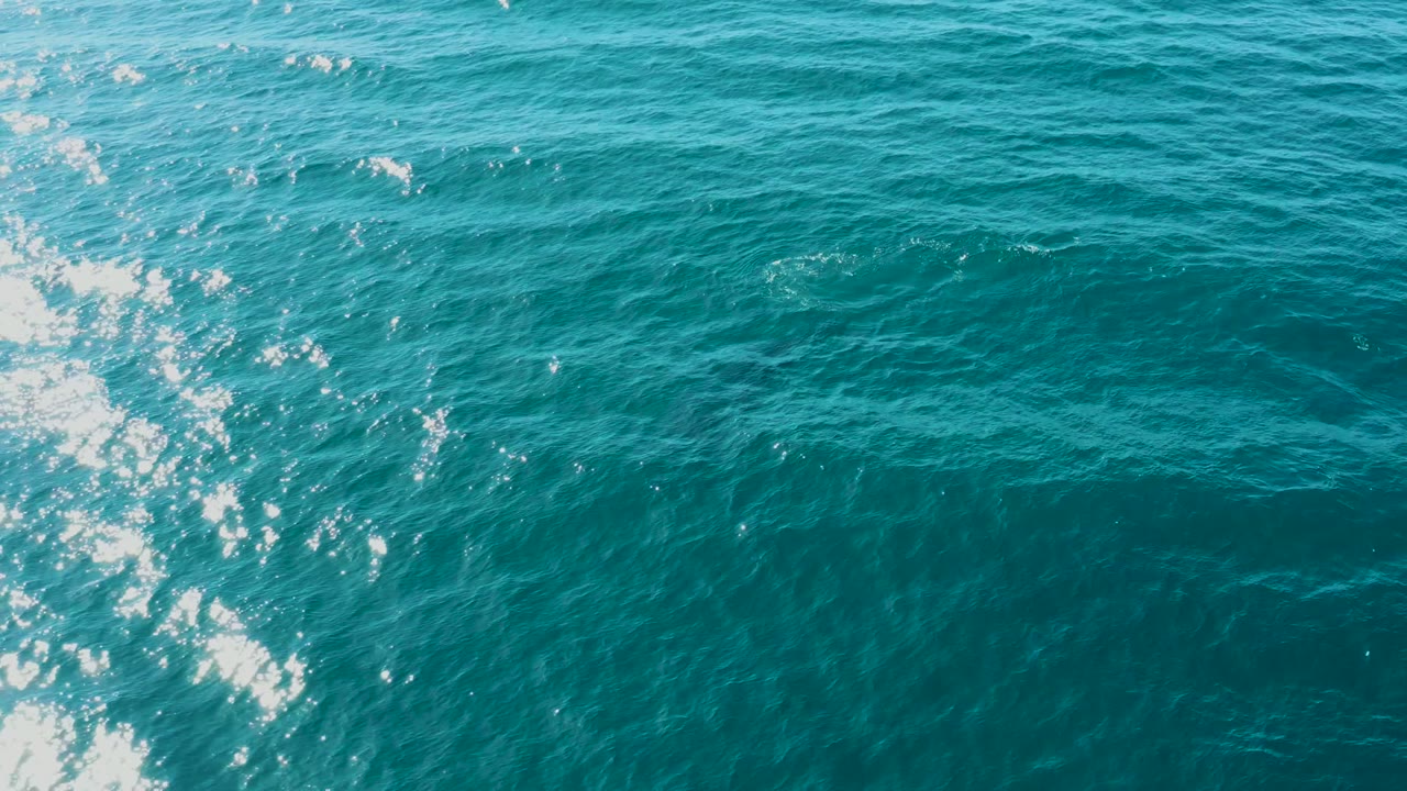 Aerial view of teal ocean at El Tule Los Cabos with a brighter vertical strip of wake foam near the left side and no visible people.