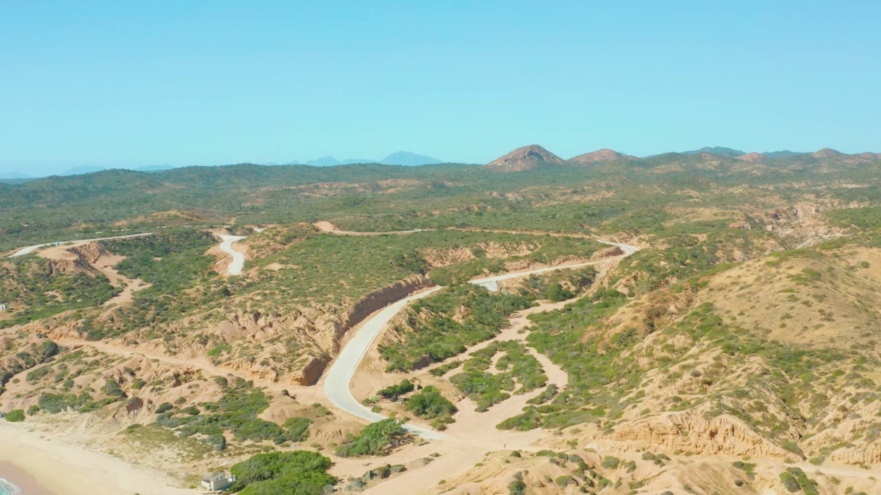 Opening aerial frame of a winding road crossing dry green hills in El Tule Los Cabos, with distant low mountains and no visible people.