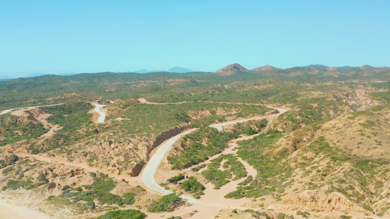 Wide elevated frame showing the winding road in the lower landscape, green-brown hills across the middle distance, and low mountains on the horizon, no visible people.