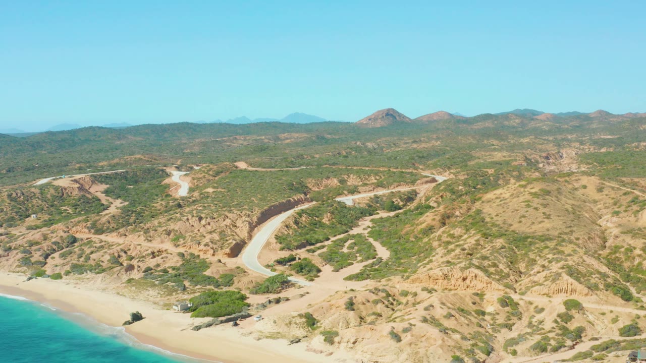 Aerial frame from El Tule Los Cabos showing a pale road curving through tan hills above a sandy beach and turquoise ocean, with no visible people.