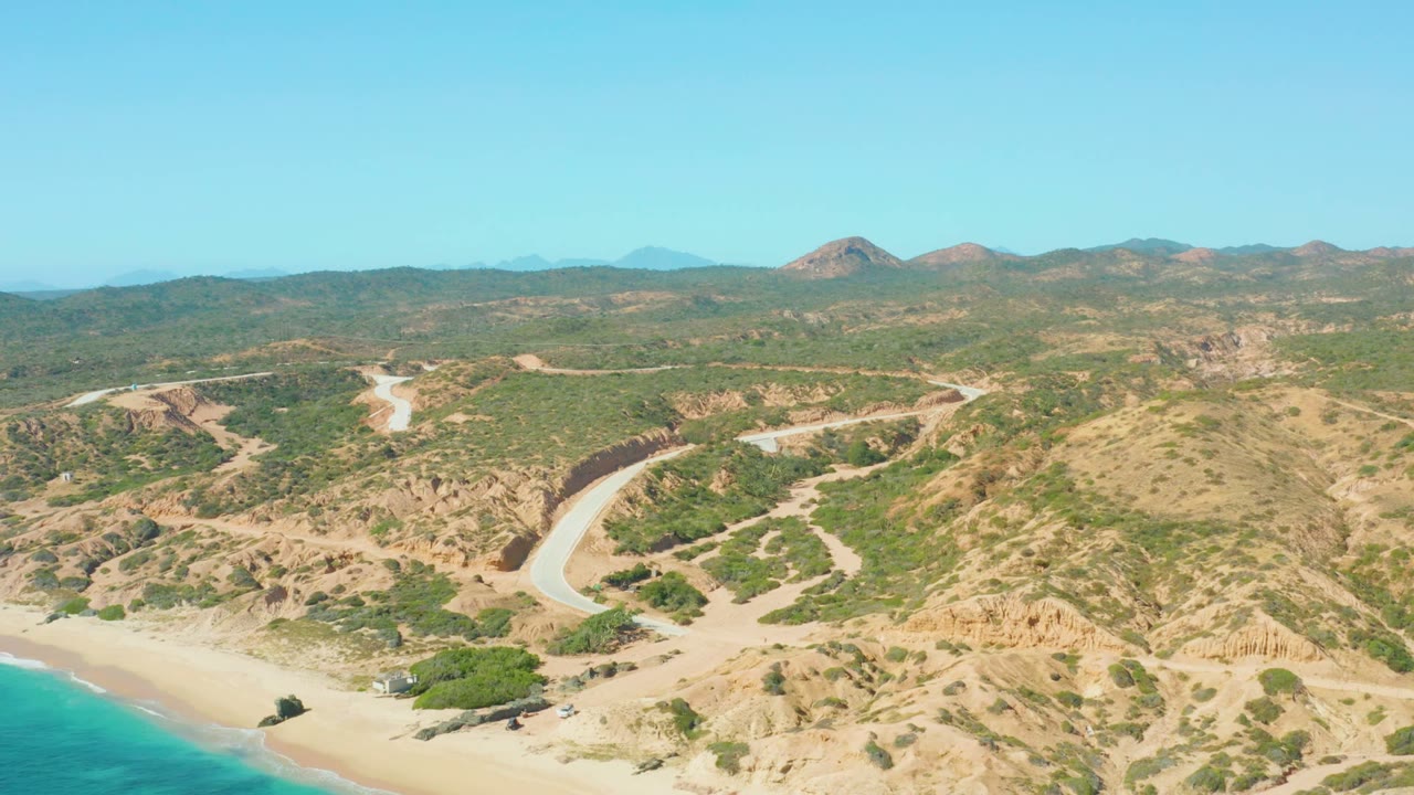Aerial view over El Tule Los Cabos with the curved road more centered between dry hills, beach sand at lower left, blue ocean water, and no visible people.