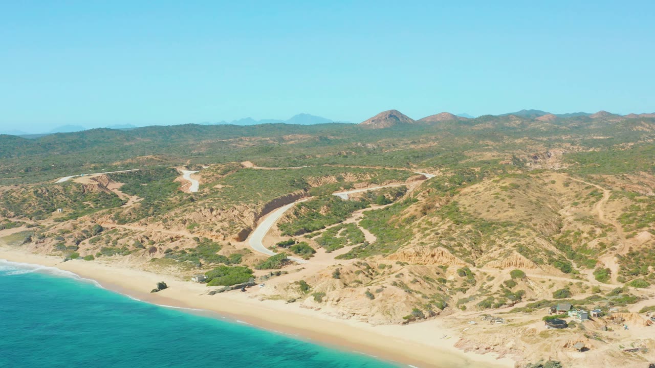 Aerial view over El Tule Los Cabos with turquoise water at lower left, sandy beach below dry hills, a pale winding road, distant mountains, and no visible people.