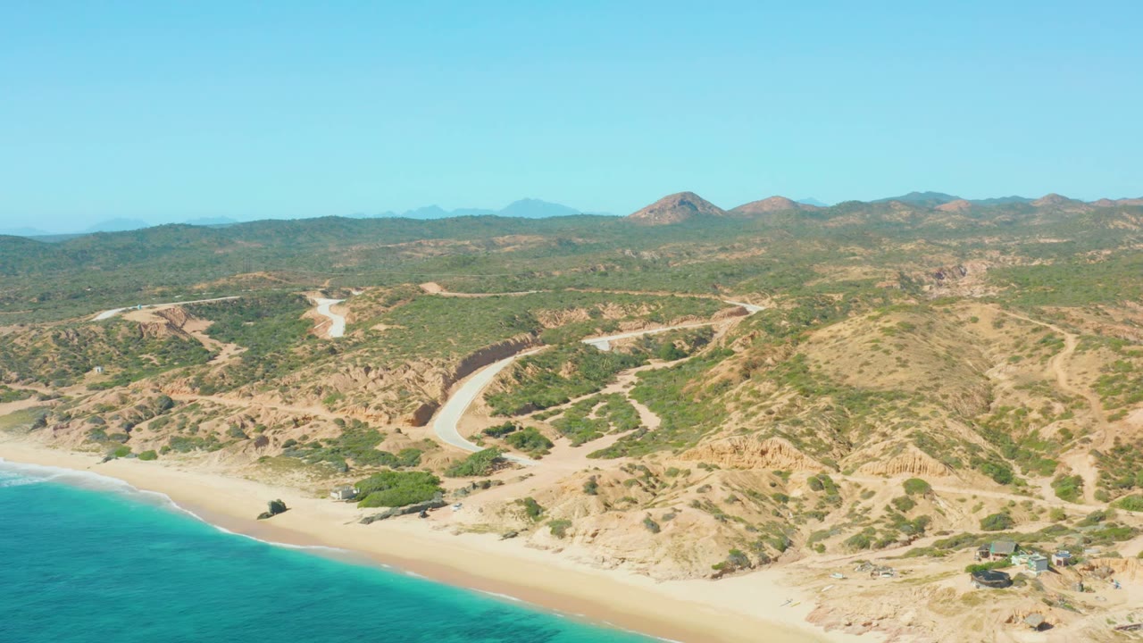 Wide aerial coastline with turquoise water, beach, scrubby hills, and pale road.