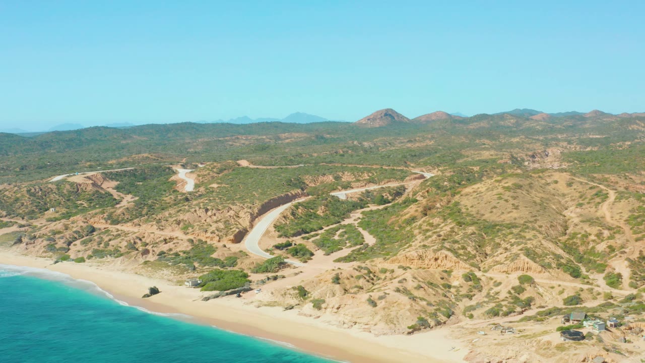 El Tule Los Cabos aerial view with the sandy beach crossing the lower frame, turquoise water at left, tan eroded hills, a road curving inland, and no visible people.