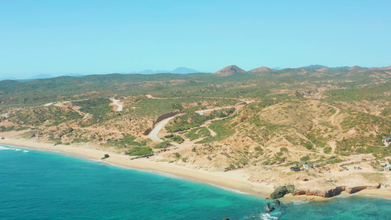 Aerial view at El Tule Los Cabos showing turquoise water along a sandy beach, arid hills behind the shore, dirt roads, small structures, and no visible people.