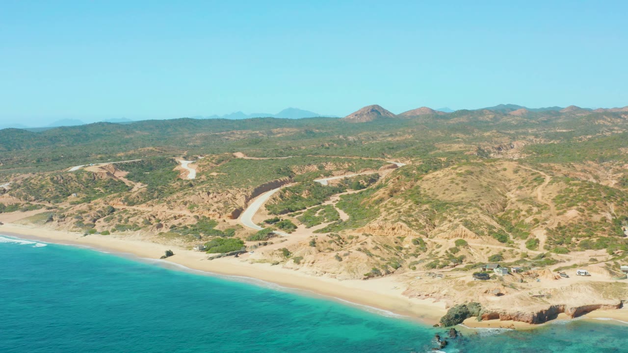 Wide aerial beach view with blue water, rocky shoreline, roads, and low hills.