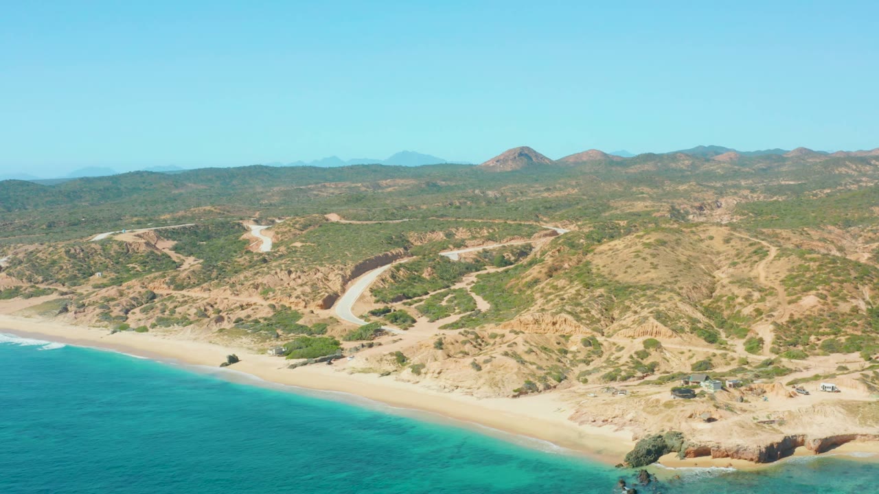 El Tule Los Cabos aerial frame with turquoise water meeting a long sandy beach, tan and green hillsides, visible road cuts, a few small vehicles or structures, and no visible people.
