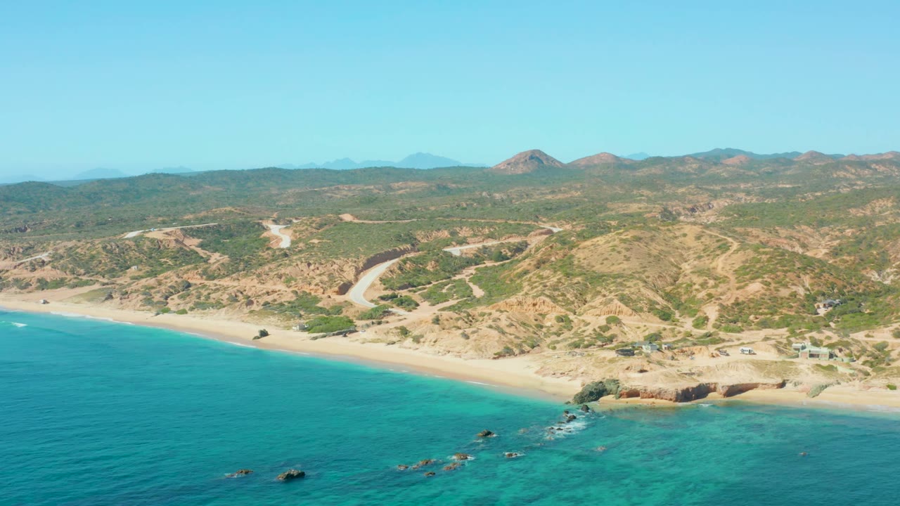 Opening aerial frame over El Tule Los Cabos, Mexico, with turquoise ocean in the foreground, a sandy beach curving below dry hills, a winding road inland, and no visible people.