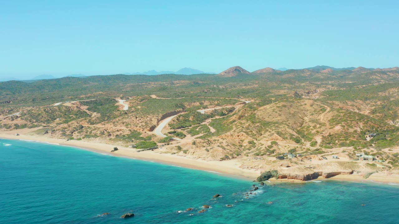 Wide coastal aerial with blue water, pale sand, rocky point, hills, and road.