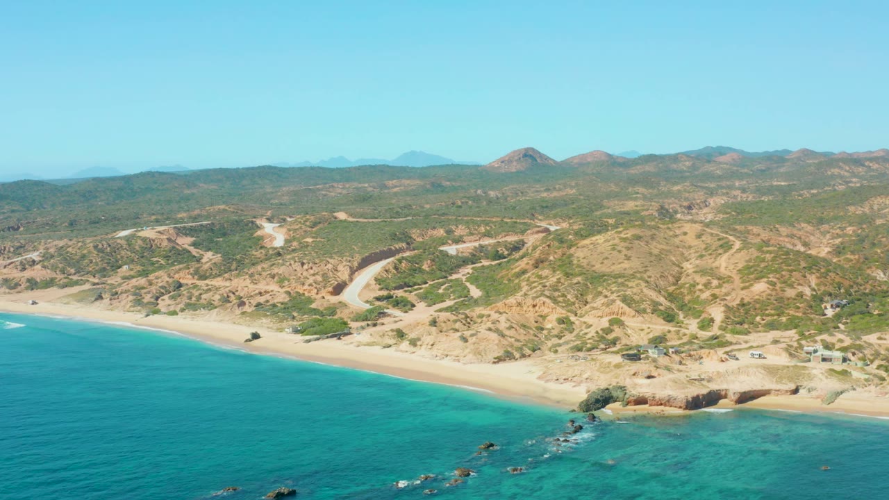 El Tule Los Cabos aerial view with turquoise water across the lower frame, sand and rocks along the shore, a winding road through dry hills, distant mountains, and no visible people.