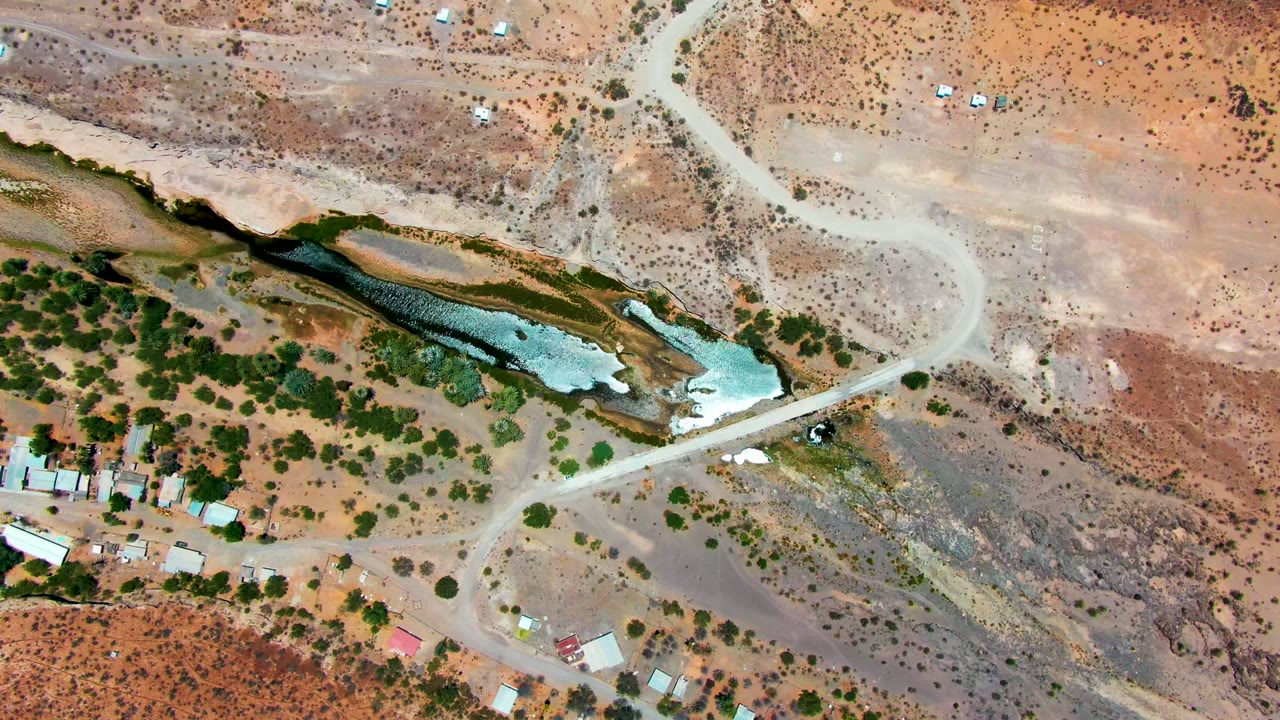 Opening overhead view in San Juanico, Baja California Sur, showing a pale dirt road crossing dry brown terrain beside a narrow greenish water body, with no visible people.