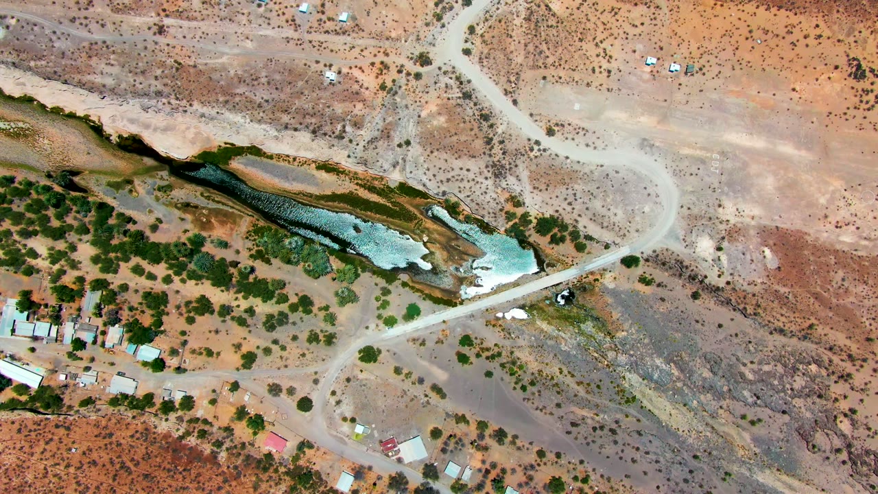 Top-down view over San Juanico, Baja California Sur, where a curved pale dirt road passes above algae-colored shallow water and scattered desert vegetation, with no visible people.
