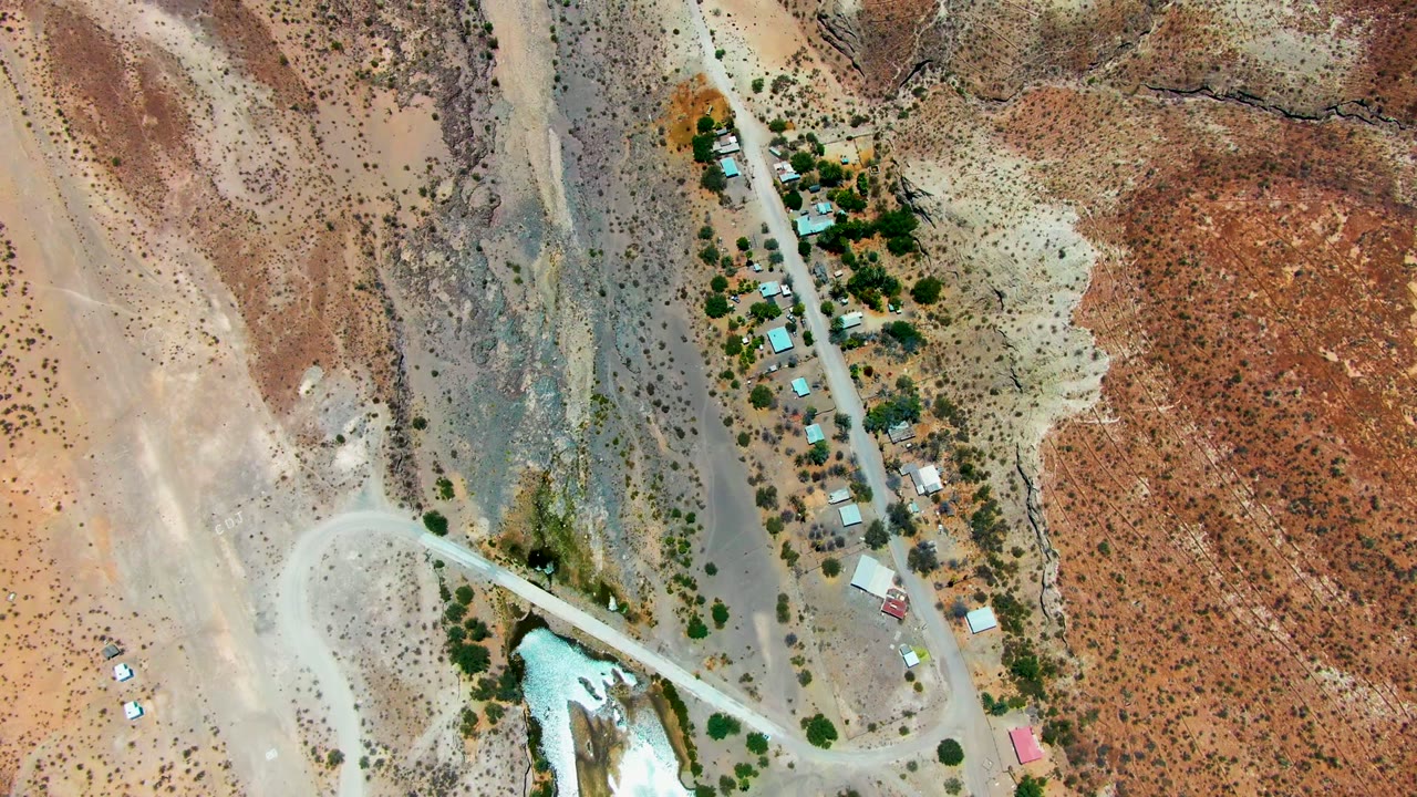 Overhead frame from San Juanico, Baja California Sur showing a pale road beside scattered buildings, trees, dry washes, and red-brown desert ground, with no visible people.