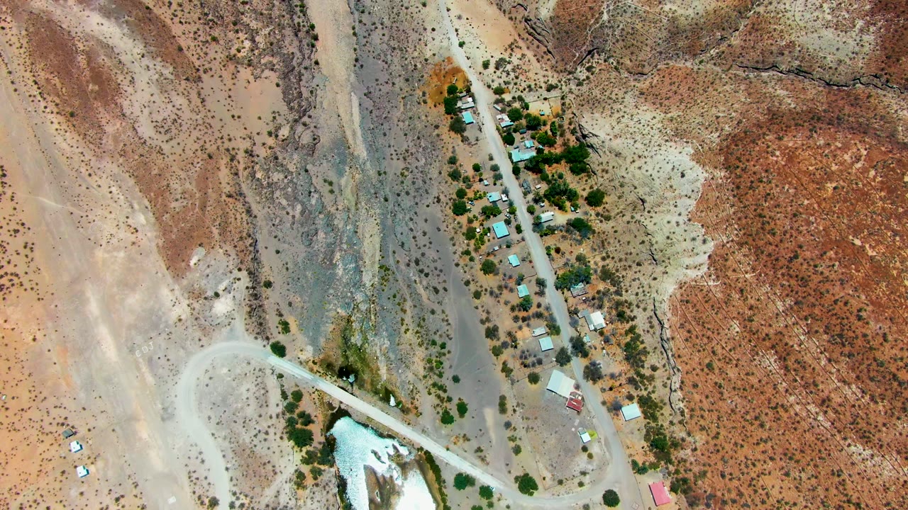 Aerial view of desert road, buildings, trees, and dry red terrain.