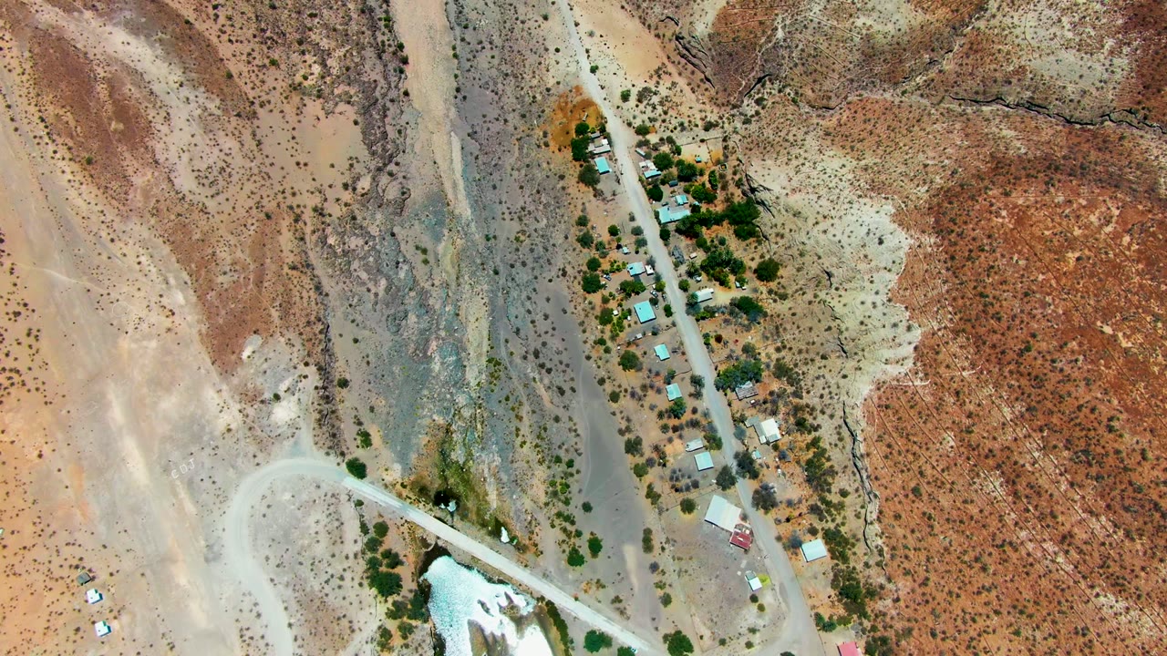 Top-down view over San Juanico, Baja California Sur showing a narrow road beside tree clusters, small structures, pale desert flats, and red-brown rocky ground, with no visible people.