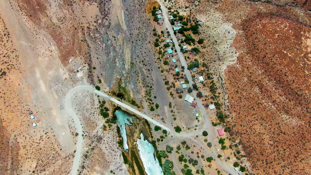 Overhead view in San Juanico, Baja California Sur of a pale road curving past a turquoise-white water channel, scattered trees, small buildings, and red desert ground with no visible people.